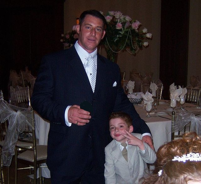 Thomas Barbusca in a grey formal suit posing with his father in a white shirt and black suit.